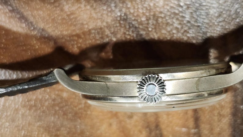 Puede incluir: Vista lateral de cerca de un reloj de pulsera plateado con una correa de cuero negro. El reloj tiene una esfera redonda y una corona decorativa.
