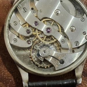 Puede incluir: Vista cercana del funcionamiento interno de un reloj de pulsera plateado con una correa de cuero negro. La esfera del reloj se ha quitado, revelando los intrincados engranajes y mecanismos.
