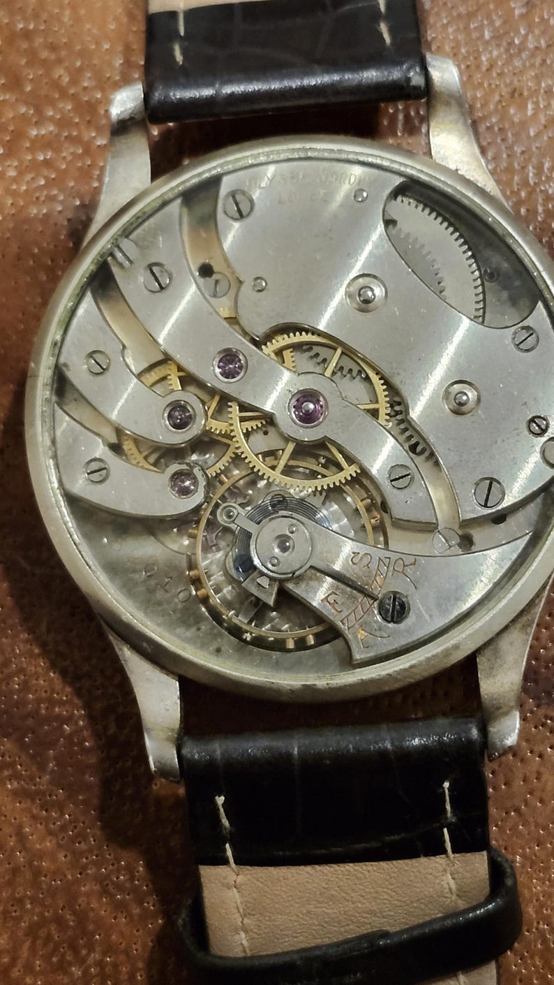 Puede incluir: Vista cercana del funcionamiento interno de un reloj de pulsera plateado con una correa de cuero negro. La esfera del reloj se ha quitado, revelando los intrincados engranajes y mecanismos.