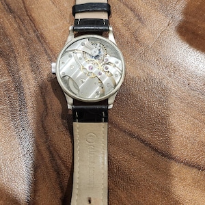 Puede incluir: Un reloj plateado con una correa de cuero negro. La esfera del reloj est&aacute; abierta, revelando el funcionamiento interno del reloj.