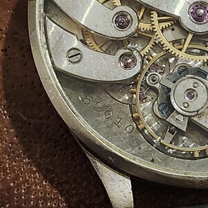Puede incluir: Un reloj plateado con una esfera transparente y una correa de cuero negro. La esfera del reloj muestra el funcionamiento interno del reloj con engranajes dorados y un fondo plateado. La esfera del reloj tiene el n&uacute;mero 52010 impreso en ella.