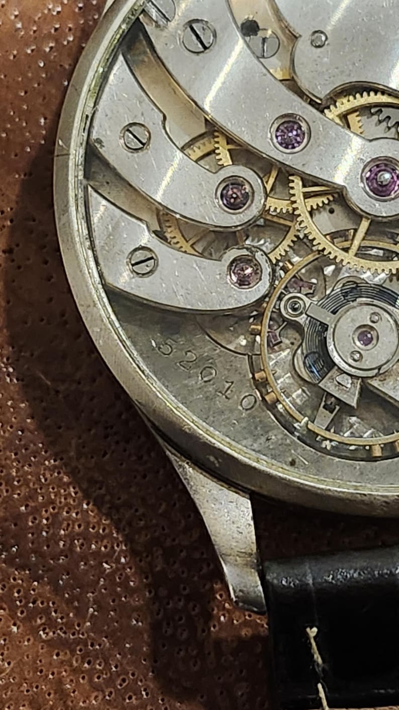 Puede incluir: Un reloj plateado con una esfera transparente y una correa de cuero negro. La esfera del reloj muestra el funcionamiento interno del reloj con engranajes dorados y un fondo plateado. La esfera del reloj tiene el n&uacute;mero 52010 impreso en ella.