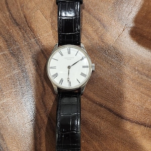 Puede incluir: Un reloj de pulsera plateado con una esfera blanca y marcadores de hora romanos negros. El reloj tiene una correa de cuero negro con un patr&oacute;n de cocodrilo.