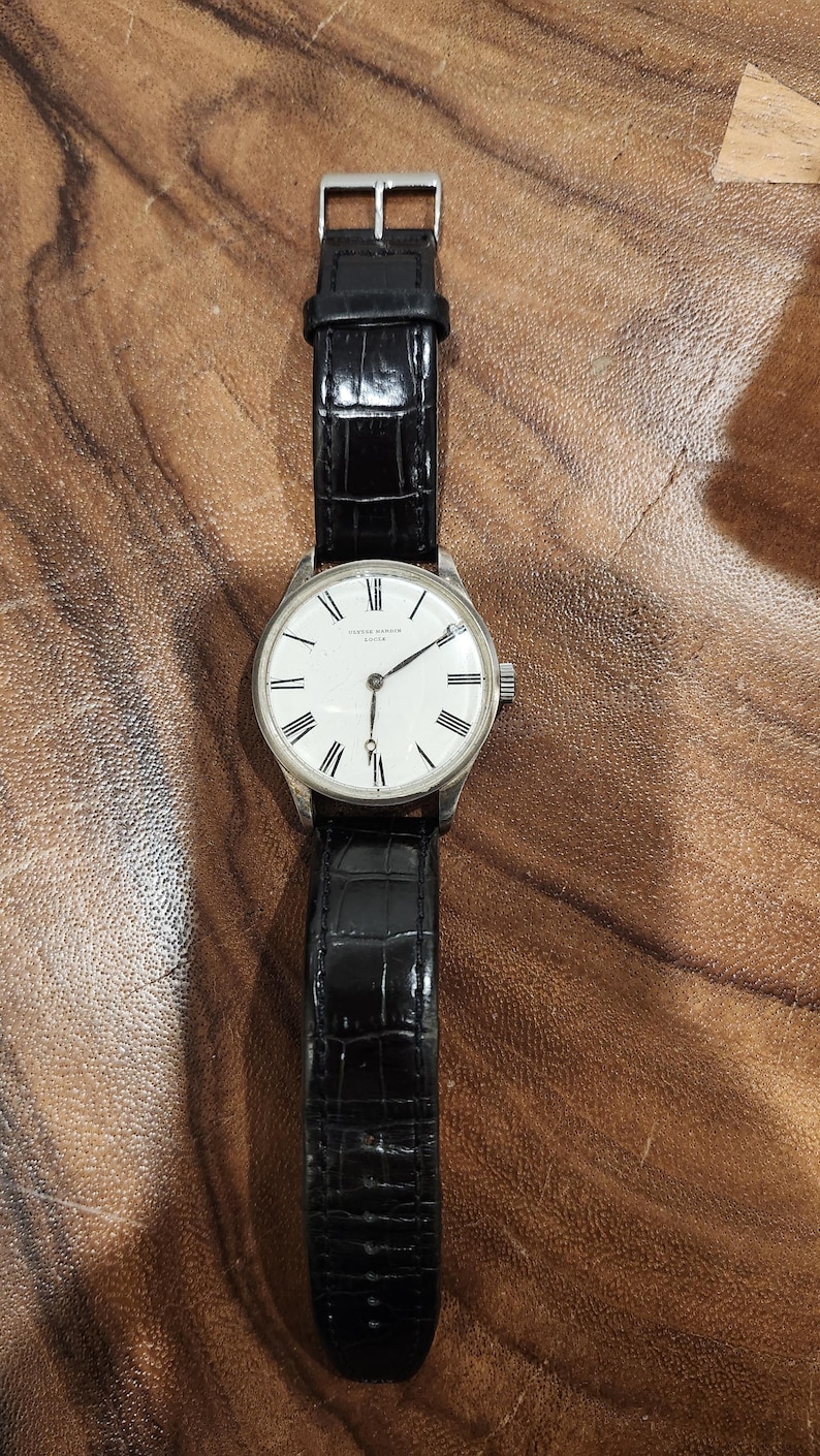 Puede incluir: Un reloj de pulsera plateado con una esfera blanca y marcadores de hora romanos negros. El reloj tiene una correa de cuero negro con un patr&oacute;n de cocodrilo.