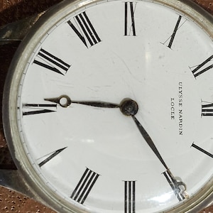 Puede incluir: Un reloj de pulsera de esfera blanca con marcadores de hora en n&uacute;meros romanos negros y agujas negras. La esfera del reloj tiene el texto "ULYSSE NARDIN LOCLE" impreso en ella. El reloj tiene una correa de cuero negro.