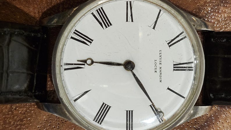 Puede incluir: Un reloj de pulsera de esfera blanca con marcadores de hora en n&uacute;meros romanos negros y agujas negras. La esfera del reloj tiene el texto "ULYSSE NARDIN LOCLE" impreso en ella. El reloj tiene una correa de cuero negro.