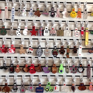 Op de afbeelding: Een wanddisplay met kleurrijke, geborduurde sleutelhangers met verschillende ontwerpen, waaronder sportteams, dieren en vakantiethema's. De sleutelhangers zijn bevestigd aan haken op een witte lattenwand.