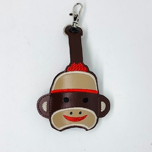 May include: A brown and tan sock monkey keychain with a red hat and a smiling face. The keychain has a silver clasp for easy attachment. The monkey's face is embroidered with a red mouth and black eyes. The keychain is made of a soft material.