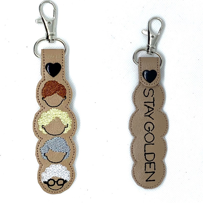 Op de afbeelding: Twee sleutelhangers met een bruine leren basis. De ene sleutelhanger heeft vier geborduurde gezichten van de Golden Girls en een zilveren gespleten ring. De andere sleutelhanger heeft de tekst "STAY GOLDEN" en een zwart hart.