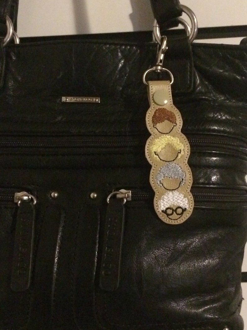 Op de afbeelding: Een zwarte leren handtas met een ritssluiting en een sleutelhanger met een stapel van vier geborduurde gezichten. De gezichten zijn bruin, blond, grijs en bruin met een bril.