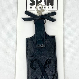 May include: Black leather keychain with a silver clasp and a black bow. The keychain has an embossed letter "X". The product is attached to a white card with the text "SPIN MARNIE crafts".