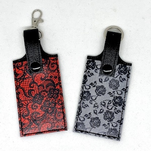 May include: Two keychains with black leather straps and metal rings. One keychain has a red background with a black lace pattern. The other keychain has a gray background with a black lace pattern.