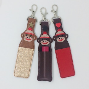 May include: Three keychains with a monkey face design. The keychains are made of brown, red, and gold leather. Each keychain has a different design on the monkey's hat: a star, a button, and a heart. The keychains are designed to hold a lipstick or lip balm.