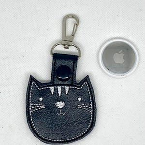 May include: Black cat-shaped keychain with white embroidered details, a silver clasp, and a black snap. An Apple AirTag is next to the keychain. The keychain is made of a textured material.