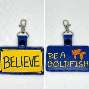 May include: Two blue leather keychains with silver clasps. One keychain has a yellow rectangle with the word "BELIEVE" embroidered in black. The other keychain has a blue rectangle with the words "BE A GOLDFISH" embroidered in white and a small orange goldfish embroidered on the right side.