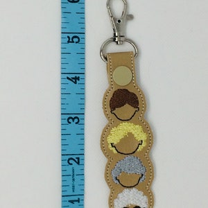 Op de afbeelding: Een beige leren sleutelhanger met een zilveren clip en vier geborduurde gezichten van vrouwen met verschillende kapsels. De sleutelhanger is ongeveer 10 cm lang.