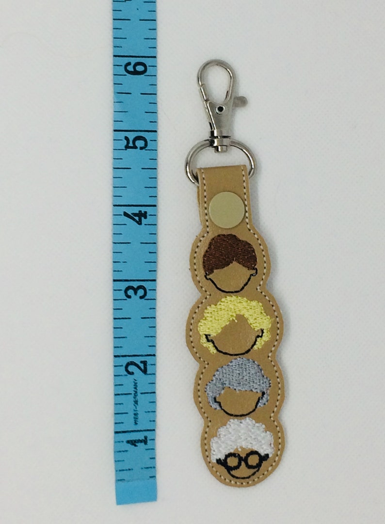 Op de afbeelding: Een beige leren sleutelhanger met een zilveren clip en vier geborduurde gezichten van vrouwen met verschillende kapsels. De sleutelhanger is ongeveer 10 cm lang.
