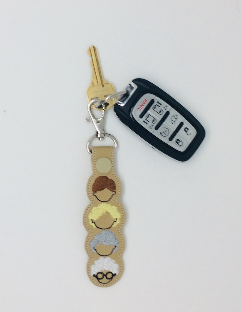 Op de afbeelding: Een beige leren sleutelhanger met vier geborduurde gezichten van vrouwen met verschillende haarkleuren en -stijlen. De sleutelhanger is bevestigd aan een zilveren sleutelring met een gouden sleutel en een zwarte autosleutel met het woord "PANIC" erop gedrukt.