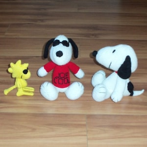 May include: Three knitted plush toys: a yellow bird with black eyes, a white dog wearing sunglasses and a red shirt with "JOE COOL" written on it, and a white dog with black ears and a black nose.