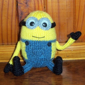 May include: A knitted Minion toy in yellow and blue. The Minion has large, round eyes with gray frames, black gloves, and black shoes. It is wearing blue overalls with black buttons and has a smiling mouth.
