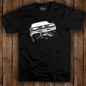 May include: Black t-shirt featuring a white graphic of a classic car with the license plate reading "RECKLESS." The car is depicted in a dynamic, lifted position. The shirt has a crew neck and short sleeves. The brand logo is visible at the top.