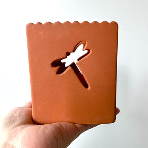 May include: A terracotta-colored ceramic planter with a scalloped edge and a dragonfly cutout. The planter is held in a hand.