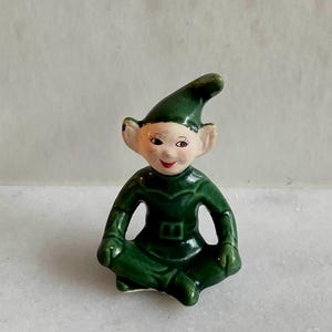 May include: A small ceramic figurine of an elf sitting cross-legged. The elf has a pale face with painted features, including dark eyes and red lips, and wears a glossy dark green outfit with a matching pointed hat and a belt detail at the waist.