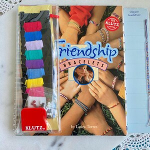 May include: A Klutz Friendship Bracelets instructional book by Laura Torres featuring a cover with multiple wrists wearing colorful woven bracelets. A clear plastic pouch attached to the side contains assorted embroidery floss, beads, and a red tool.