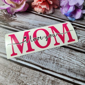 May include: A white vinyl sticker with the word "MOM" in large pink letters. The words "I love you" are written in black cursive script above the word "MOM" with a small heart symbol.