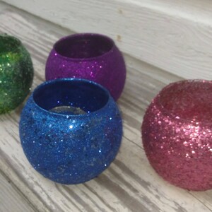 May include: Four glitter-covered, spherical candle holders in green, purple, blue, and pink. The holders are open at the top and sit on a light-colored wooden surface. The glitter adds a sparkling, festive touch.