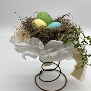 Könnte beinhalten: Osterdekoration mit einem Nest mit drei gesprenkelten Eiern in Grün, Gelb und Blau. Das Nest besteht aus natürlichen Materialien und sitzt auf einer weißen Blume und einer rostigen Metallfederbasis. Ein frühlingshaftes Dekorationsstück.