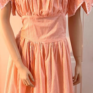 May include: A vintage-style dress in a peach and white gingham pattern. The dress features a square neckline, short sleeves, and a fitted waist. White lace trim accents the neckline, sleeves, and hem. The dress is displayed on a mannequin.