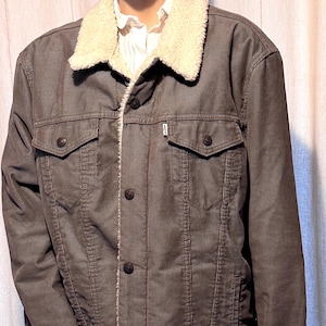 May include: A brown corduroy jacket with a cream-colored sherpa collar and lining. The jacket has two chest pockets, two side pockets, and button closures. The jacket is displayed on a mannequin.