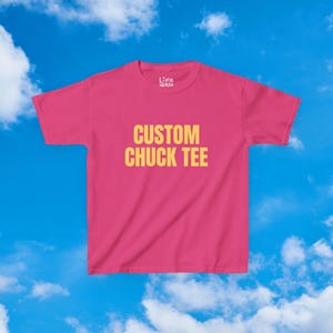 Pode incluir: Uma t-shirt de manga curta e gola redonda numa cor rosa vibrante. O centro do peito apresenta o texto "CUSTOM CHUCK TEE" em letras maiúsculas amarelas, a negrito e sem serifas. A etiqueta interior da gola exibe o texto "LITTLE WEIRDO".