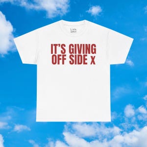 Puede incluir: Camiseta blanca con el texto "IT'S GIVING OFF SIDE X" en rojo. La camiseta tiene cuello redondo y mangas cortas. La marca "Little Weirdo" está impresa en la parte superior de la camiseta. El fondo es un cielo azul con nubes blancas.