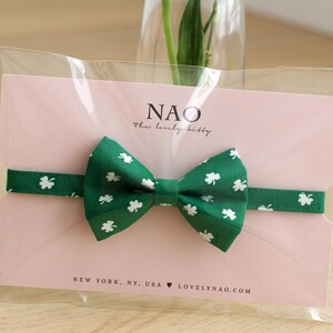 May include: A green bow tie with white shamrock print, packaged in a clear plastic bag with a pink card that reads "NAO the lovely kitty" and "NEW YORK, NY, USA LOVELYNAO.COM".