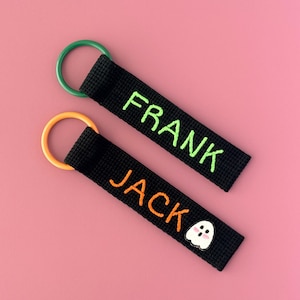 May include: Two black fabric keychains with circular rings. One keychain has a green ring and the name FRANK embroidered in green thread. The other has an orange ring, the name JACK in orange thread, and a small white ghost icon with pink cheeks.