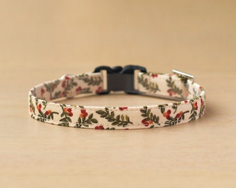 May include: A white fabric dog collar with a floral pattern of green leaves and red berries. The collar has a black plastic buckle.