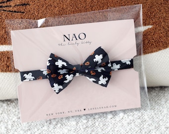 May include: A black bow tie with a white ghost and orange pumpkin pattern. The bow tie is packaged in a clear plastic bag with a pink card that says "NAO the lovely kitty" and "NEW YORK, NY, USA LOVELYNAO.COM".