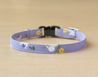 以下が含まれることがあります： 白いうさぎと黄色の花の模様がついたラベンダー色の猫用首輪。首輪には黒いプラスチック製のバックルと銀色の金属製のDリングが付いています。