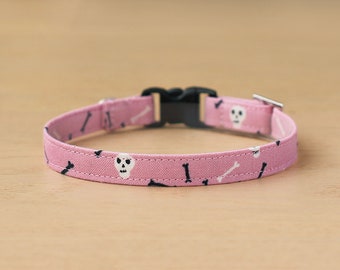 May include: Pink fabric pet collar with a black buckle and a pattern of white skulls and black bones.