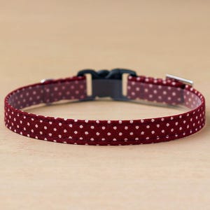 May include: A burgundy polka dot collar with a black plastic buckle and a silver metal clasp. The collar is made of fabric and features small white polka dots. The collar is designed for a pet.