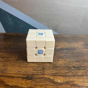 May include: A cream-colored Rubik's Cube with blue and white logos on each square. The cube is sitting on a dark brown wooden surface. The background features a wall with a geometric design.