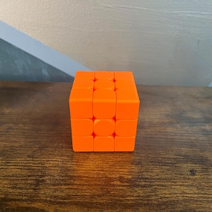 May include: An orange Rubik's Cube sits on a dark wooden surface. The cube is a 3x3 puzzle, with each side composed of nine squares. The cube's bright orange color contrasts with the wood grain.