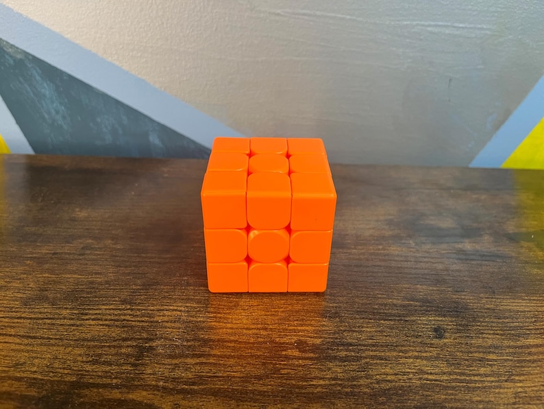 May include: An orange Rubik's Cube sits on a dark wooden surface. The cube is a 3x3 puzzle, with each side composed of nine squares. The cube's bright orange color contrasts with the wood grain.