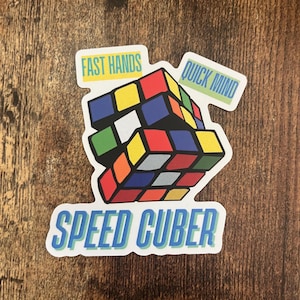 May include: A sticker featuring a colorful Rubik's Cube with the phrases "Fast Hands" and "Quick Mind" on separate labels. The sticker also includes the text "Speed Cuber" in blue and green.