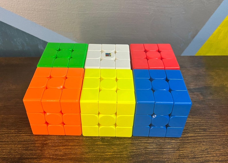 May include: A collection of six multi-colored Rubik's Cubes, each with a 3x3 grid of squares. The colors include green, orange, yellow, white, red, and blue. The cubes are arranged in a rectangular shape.