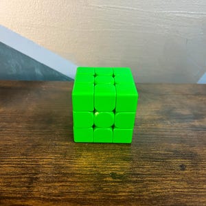 May include: A bright green Rubik's Cube sits on a dark wood surface. The cube is a classic puzzle toy with a 3x3 grid of colored squares. The background features a wall with geometric shapes in shades of gray, blue, and yellow.