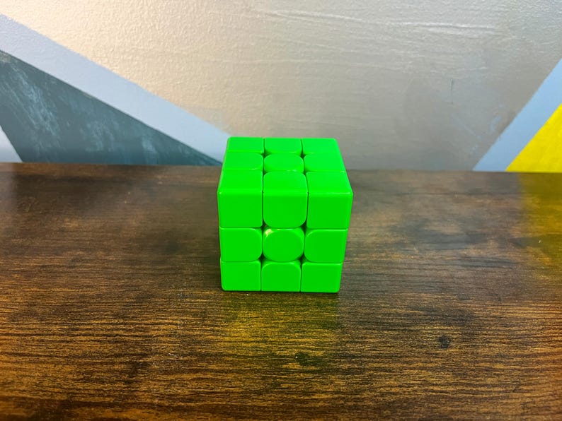 May include: A bright green Rubik's Cube sits on a dark wood surface. The cube is a classic puzzle toy with a 3x3 grid of colored squares. The background features a wall with geometric shapes in shades of gray, blue, and yellow.