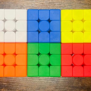 May include: Six colorful Rubik's Cubes arranged in two rows of three. Each cube is composed of nine squares in a 3x3 grid. The colors are white, blue, yellow, orange, green, and red. The cubes are on a wooden surface.